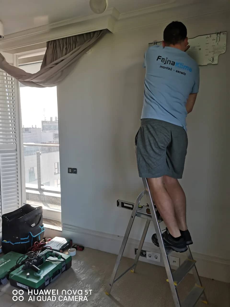 Monter w koszulce FajnaKlima na drabinie przy ścianie apartamentu z zasłonami i okiennicami