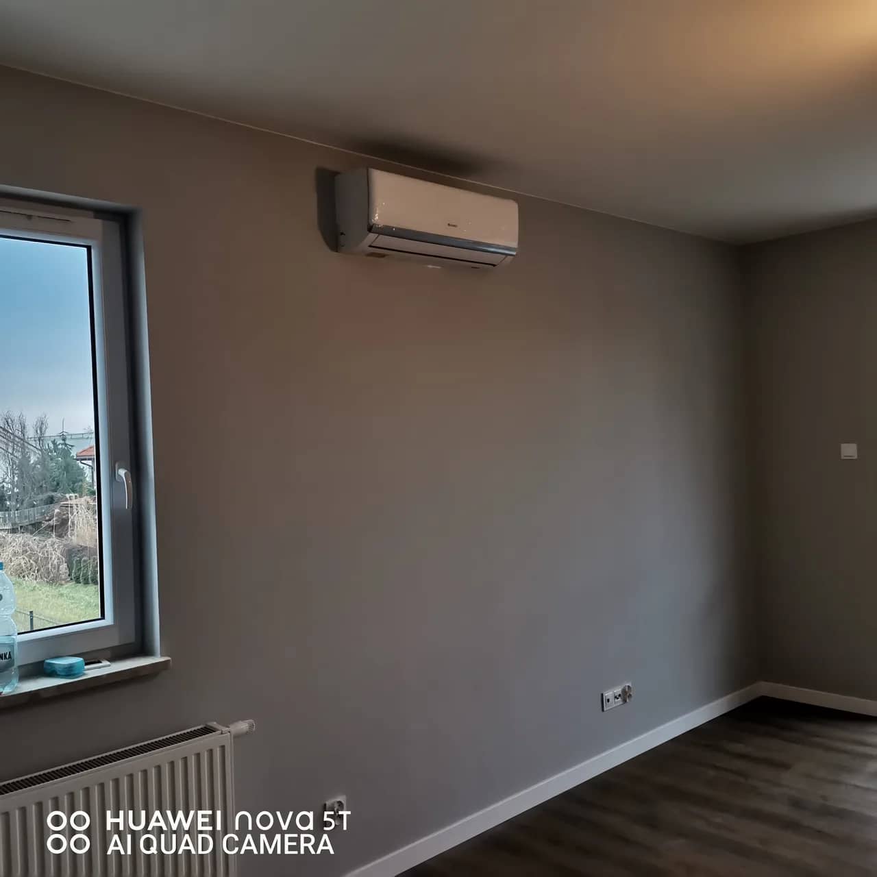 Biały klimatyzator ścienny na szarej ścianie pokoju z podłogą z desek i grzejnikiem