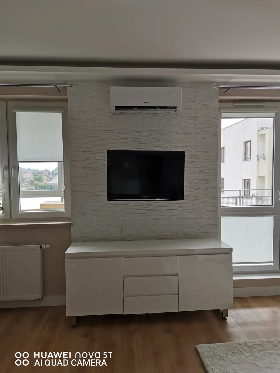 Salon z telewizorem na kamiennej ścianie, białą komodą i klimatyzatorem pod sufitem