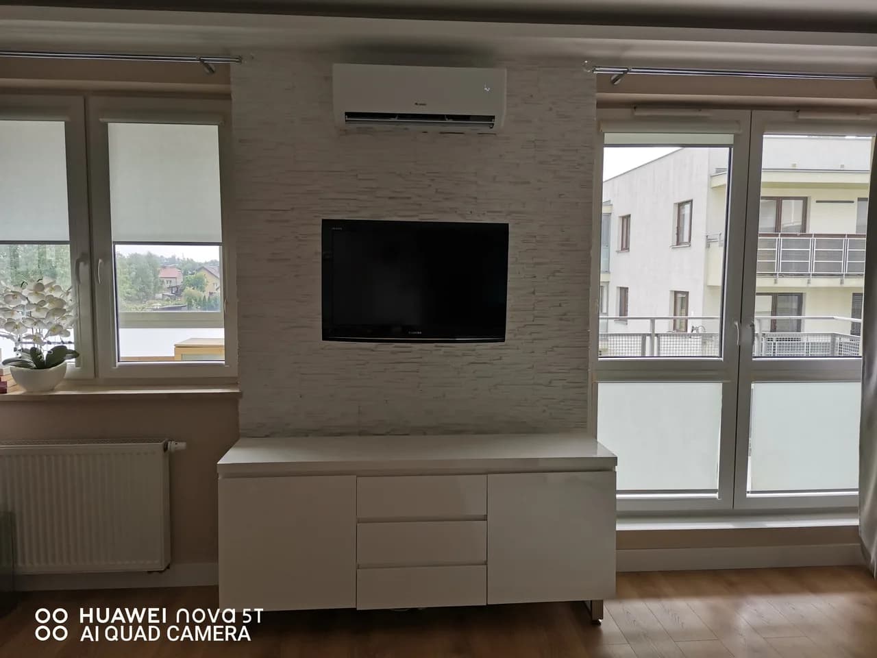 Klimatyzator ścienny widziany pod kątem, obok okna i grzejnika w jasnym salonie