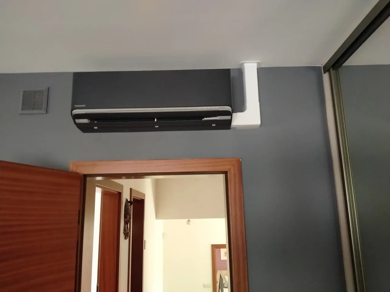 Grafitowa jednostka wewnętrzna klimatyzacji na szarej ścianie poddasza, obok otwarte drzwi