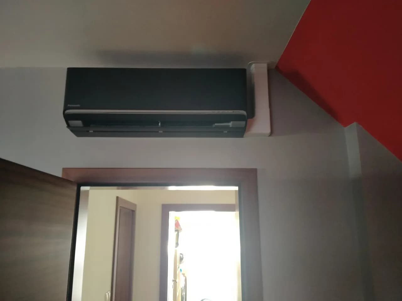 Klimatyzator ścienny w rogu pokoju z czerwoną ścianą i widokiem na korytarz