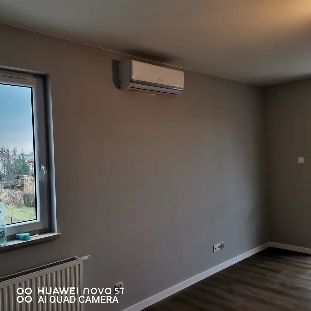 Biały klimatyzator ścienny na szarej ścianie pokoju z podłogą z desek i grzejnikiem